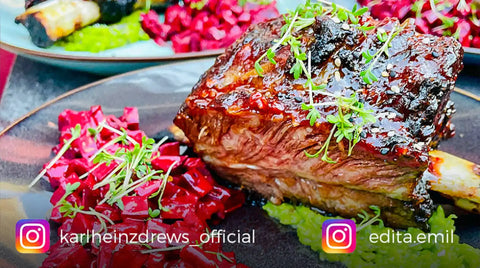 Ein Teller mit glasierten AUMAERK-Rindsrippen, garniert mit frischen Kräutern, serviert mit lebhaftem Rotkohl und einem grünen Püree. Ein anderer Teller mit ähnlichen Speisen ist im Hintergrund verschwommen. Die Griffe von Instagram erscheinen unten.