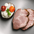 Zwei Scheiben AUMAERK-Schweinsbraten vom Schopf „Noble Roast Cut“ liegen neben einer Scheibe Brot, die mit Ricotta, halbierten Kirschtomaten, Mozzarella-Kugeln und frischem Basilikum belegt ist, serviert auf einem Silberteller.