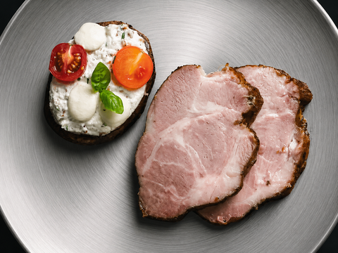 Zwei Scheiben AUMAERK-Schweinsbraten vom Schopf „Noble Roast Cut“ liegen neben einer Scheibe Brot, die mit Ricotta, halbierten Kirschtomaten, Mozzarella-Kugeln und frischem Basilikum belegt ist, serviert auf einem Silberteller.
