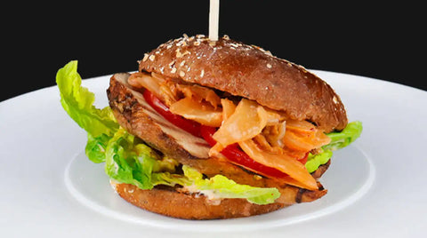 Ein Sandwich mit gegrilltem AUMAERK-Hähnchen, Salat, Tomate und eingelegtem Gemüse auf einem Vollkornbrötchen, serviert auf einem weißen Teller vor schwarzem Hintergrund.