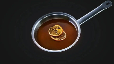 Ein AUMAERK-Metalltopf mit Henkel enthält eine dunkelbraune Flüssigkeit, auf der zwei getrocknete Orangenscheiben vor einem schwarzen Hintergrund liegen.