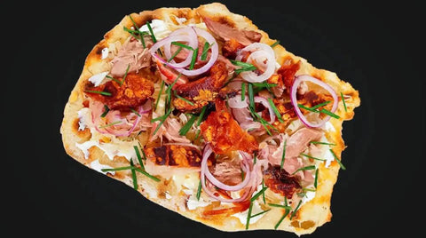 Ein unregelmäßig geformtes Fladenbrot, belegt mit AUMAERK-Hackfleisch, geschnittenen roten Zwiebeln, Schnittlauch und geschmolzenem Käse auf schwarzem Hintergrund.