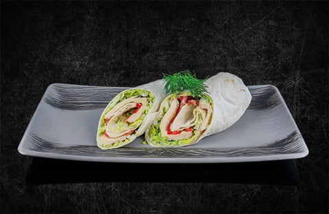 Ein sauber arrangiertes, halbiertes AUMAERK-Wrap-Sandwich auf einem rechteckigen grauen Teller, gefüllt mit Grünzeug, roter Paprika und Delikatessfleisch, garniert mit einem frischen Dillzweig, vor einem dunklen, strukturierten Hintergrund.