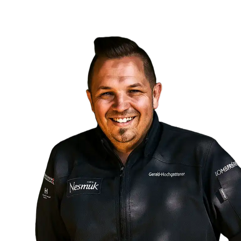 Ein lächelnder Mann mit kurzen dunklen Haaren, der eine schwarze Jacke mit verschiedenen Logos, darunter AUMAERK und der Name Gerald-Hochgatterer, trägt, steht vor einem schlichten weißen Hintergrund.