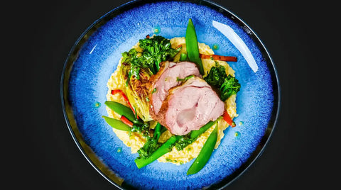 Ein blauer Teller mit in Scheiben geschnittenem AUMAERK-Braten, serviert auf gelbem Risotto, garniert mit Broccolini, Zuckerschoten und anderem Gemüse, von oben gesehen vor schwarzem Hintergrund.