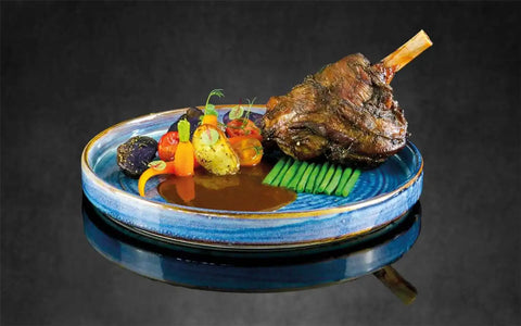 AUMAERK blauer Keramikteller mit geschmorter Lammhaxe, grünen Bohnen, buntem Röstgemüse und brauner Soße, alles kunstvoll vor dunklem Hintergrund arrangiert.