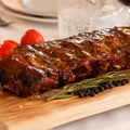 AUMAERKs Spareribs „Little Rocco“ – Limited Edition, glasiert und auf einem Holzbrett gegrillt, garniert mit Rosmarin, Pfefferkörnern, Knoblauch und Kirschtomaten. Elegantes Geschirr und eine brennende Kerze sorgen im Hintergrund für eine warme Atmosphäre.