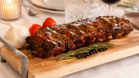 AUMAERKs Spareribs „Little Rocco“ – Limited Edition, glasiert und auf einem Holzbrett gegrillt, garniert mit Rosmarin, Pfefferkörnern, Knoblauch und Kirschtomaten. Elegantes Geschirr und eine brennende Kerze sorgen im Hintergrund für eine warme Atmosphäre.