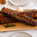 Die Spareribs „Little Rocco“ von AUMAERK in limitierter Auflage werden auf einem Holzbrett serviert und mit Kräutern, Pfefferkörnern, Knoblauch, roten Chilischoten und grobem Salz garniert – perfekt für eine elegante Tischdekoration mit Wein und Kerzen.