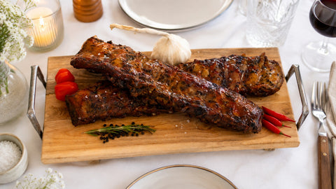 Die Spareribs „Little Rocco“ von AUMAERK in limitierter Auflage werden auf einem Holzbrett serviert und mit Kräutern, Pfefferkörnern, Knoblauch, roten Chilischoten und grobem Salz garniert – perfekt für eine elegante Tischdekoration mit Wein und Kerzen.