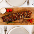 Die Spareribs „Little Rocco“ von AUMAERK – Limited Edition, mit BBQ-Glasur und garniert mit Kräutern, Knoblauch und roten Chilischoten – werden auf einem Brett an einem Tisch serviert, der mit Tellern, Gläsern, Besteck und einer Stoffserviette gedeckt ist.