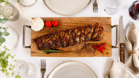 Die Spareribs „Little Rocco“ von AUMAERK – Limited Edition, mit BBQ-Glasur und garniert mit Kräutern, Knoblauch und roten Chilischoten – werden auf einem Brett an einem Tisch serviert, der mit Tellern, Gläsern, Besteck und einer Stoffserviette gedeckt ist.