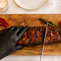 Eine Hand in einem schwarzen Handschuh schneidet AUMAERKS Spareribs „Little Rocco“ aus der Limited Edition auf einem Holzbrett, umgeben von roten Chilischoten, Kräutern, einer Kerze, Wein und einem gedeckten Tisch.