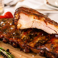 Die Spareribs „Little Rocco“ von AUMAERK – Limited Edition – werden auf einem Holzbrett serviert, mit grobem Salz bestreut und mit Rosmarin, schwarzen Pfefferkörnern, Tomaten und Knoblauch im Hintergrund garniert.