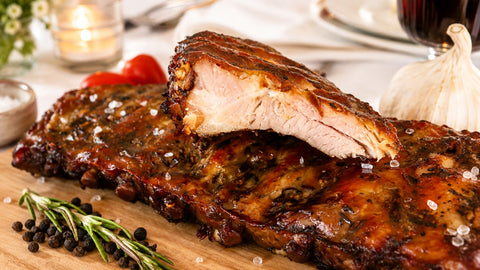 Die Spareribs „Little Rocco“ von AUMAERK – Limited Edition – werden auf einem Holzbrett serviert, mit grobem Salz bestreut und mit Rosmarin, schwarzen Pfefferkörnern, Tomaten und Knoblauch im Hintergrund garniert.