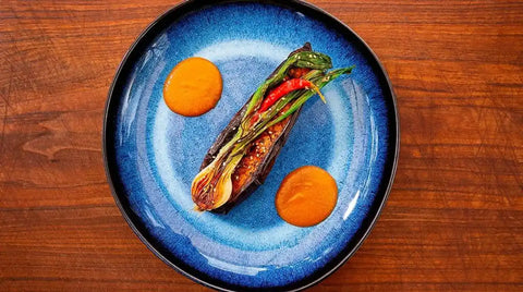 Ein blauer Teller mit einer gebratenen Aubergine, garniert mit grünen Bohnen, rotem Chili und anderem Gemüse von AUMAERK, garniert mit zwei orangefarbenen Soßentupfern, steht auf einem Holztisch.