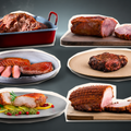 Die „Royal Six – Gourmet-Probierbox“ von AUMAERK enthält fünf erlesene Fleischspezialitäten: Braten, serviert in einer roten Pfanne, aufgeschnittenen Braten, präsentiert auf einem Brett, eine Auswahl an Würsten und Fleischsorten aus der „Royal Six“-Auswahl, ein perfekt gegrilltes Steak sowie Schinkenscheiben auf eleganten Tellern – alles kunstvoll auf einem dunklen Hintergrund angerichtet.