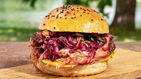 Ein AUMAERK Pulled Pork-Sandwich auf einem entkernten Brötchen, gefüllt mit gelbem Käse und belegt mit kräftigem Rotkohlsalat, liegt auf einem Holzbrett im Freien, während die Bäume im Hintergrund verschwimmen.
