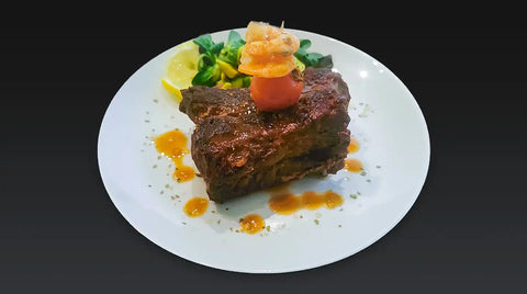 Ein weißer Teller mit einer Portion gegrilltem AUMAERK-Fleisch, belegt mit einer Kirschtomate und einer Beilage, beträufelt mit Sauce, begleitet von Blattgemüse und einer Zitronenscheibe, vor einem dunklen Hintergrund.