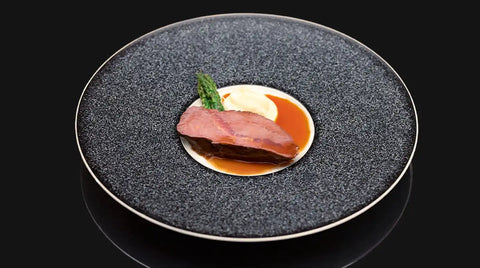 Ein Gourmet-Gericht von AUMAERK mit einer Scheibe gebratenem Fleisch, grünem Spargel, Kartoffelpüree und einer braunen Soße, kunstvoll arrangiert auf einem strukturierten schwarzen Teller vor einem schwarzen Hintergrund.