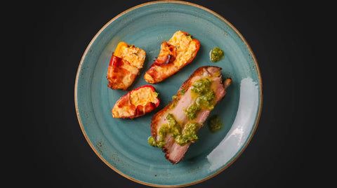 Ein blaugrüner Teller mit einer Scheibe gebratenem AUMAERK-Fleisch mit grüner Soße, serviert mit drei gefüllten, gebackenen Kartoffelhälften mit goldenem Käsebelag. Der Hintergrund ist schwarz.