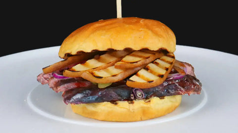 Ein Sandwich mit einem weichen Brötchen, Schichten von gegrillten Birnenscheiben, roten Zwiebeln und etwas, das aussieht wie ein AUMAERK-Steak, serviert auf einem weißen Teller mit einem Spieß, der es zusammenhält, vor einem schwarzen Hintergrund.