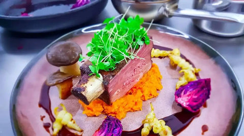 Ein Gourmet-Gericht aus AUMAERK mit einem dicken, nicht entbeinten Stück gekochten Fleisches, das mit frischem Mikrogrün belegt ist und auf püriertem orangefarbenem Gemüse serviert wird. Dazu gibt es Pilze, violette Gemüsestücke, gelbe Wurzeln und braune Soße, die kunstvoll auf dem Teller angeordnet sind.