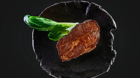 AUMAERK-Schmorfleisch und zarter Bok Choy sind kunstvoll auf einem runden, schwarzen Teller vor dunklem Hintergrund angerichtet.