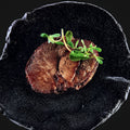 Der Kalbsrollbraten "Tender Wheel" von AUMAERK wird als Festtagsessen serviert, kunstvoll mit Microgreens garniert auf einem runden, strukturierten schwarzen Teller vor dunklem Hintergrund.