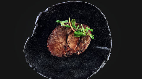 Der Kalbsrollbraten "Tender Wheel" von AUMAERK wird als Festtagsessen serviert, kunstvoll mit Microgreens garniert auf einem runden, strukturierten schwarzen Teller vor dunklem Hintergrund.