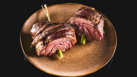 Zwei Stück vom Kalbsrollbraten "Tender Wheel" von AUMAERK, gerollt und mit grünem Gemüse gefüllt, serviert auf braunem Keramikteller vor schwarzem Hintergrund - ein besonderes Festtagsessen.