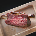 Eine Nahaufnahme einer dicken, saftigen, mittelgroßen AUMAERK-Rinderrippe, halbiert, mit rosa Kern und gegrillter Außenkruste, serviert auf einem Holztablett.
