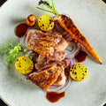 Ein Gourmet-Gericht mit Stelze "Crack Leg" von AUMAERK mit gegrillter Karotte, gelben Medaillons, roten Zwiebelringen, grüner Garnitur und brauner Soße, kunstvoll arrangiert auf einem großen weißen Teller nach der raffinierten AUMAERK-Methode.