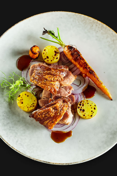 Ein Gourmet-Gericht mit Stelze "Crack Leg" von AUMAERK mit gegrillter Karotte, gelben Medaillons, roten Zwiebelringen, grüner Garnitur und brauner Soße, kunstvoll arrangiert auf einem großen weißen Teller nach der raffinierten AUMAERK-Methode.