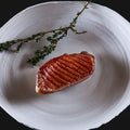 AUMAERKs Entenbrust aus "The Duke Filet Sixpack" wird mit Grillspuren gebraten, auf einem weißen Teller serviert und mit frischem Thymian vor schwarzem Hintergrund garniert.