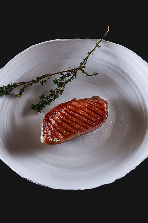AUMAERKs Entenbrust aus "The Duke Filet Sixpack" wird mit Grillspuren gebraten, auf einem weißen Teller serviert und mit frischem Thymian vor schwarzem Hintergrund garniert.