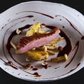 Ein Gourmet-Gericht mit gebratener Entenbrust im "The Duke Filet Sixpack" von AUMAERK, serviert mit gerösteten Kartoffeln, Pilzen und Haselnüssen, kunstvoll präsentiert mit einer dunklen Sauce auf einem weißen Teller vor schwarzem Hintergrund.