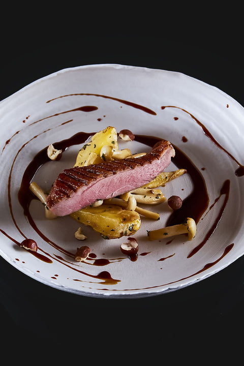 Ein Gourmet-Gericht mit gebratener Entenbrust im "The Duke Filet Sixpack" von AUMAERK, serviert mit gerösteten Kartoffeln, Pilzen und Haselnüssen, kunstvoll präsentiert mit einer dunklen Sauce auf einem weißen Teller vor schwarzem Hintergrund.