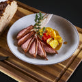 Geschnittene Entenbrust im "The Duke Filet Sixpack" von AUMAERK, elegant angerichtet mit gelbem und rotem Gemüse, frischen Kräutern und rustikalem Brot auf einem Holzbrett - ein Premium-Geflügelgericht für echte Genießer.