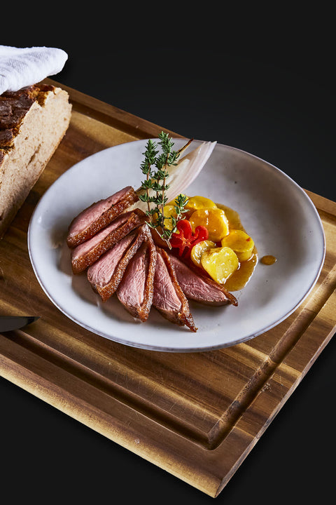 Geschnittene Entenbrust im "The Duke Filet Sixpack" von AUMAERK, elegant angerichtet mit gelbem und rotem Gemüse, frischen Kräutern und rustikalem Brot auf einem Holzbrett - ein Premium-Geflügelgericht für echte Genießer.