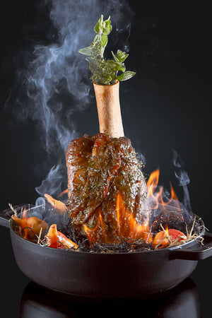 Ein AUMAERK-gekochter Braten mit Knochen steht aufrecht in einer schwarzen Pfanne mit Kräutern darauf, umgeben von Gemüse und Flammen, wobei Rauch vor einem dunklen Hintergrund aufsteigt.