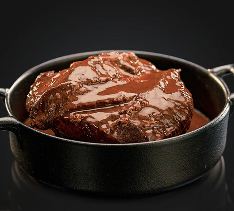 Ein dickes Stück AUMAERK-Rindfleisch, bedeckt mit einer glänzenden braunen Sauce, liegt in einer schwarzen gusseisernen Pfanne vor einem dunklen Hintergrund.