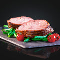 Zwei dicke Scheiben Kalbsrollbraten "Tender Wheel" von AUMAERK, serviert auf grünen Bohnen und garniert mit ganzen Cherrytomaten - ein festliches Gericht auf dunkler Schieferplatte vor schwarzem Hintergrund.