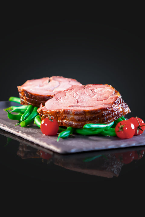 Zwei dicke Scheiben Kalbsrollbraten "Tender Wheel" von AUMAERK, serviert auf grünen Bohnen und garniert mit ganzen Cherrytomaten - ein festliches Gericht auf dunkler Schieferplatte vor schwarzem Hintergrund.