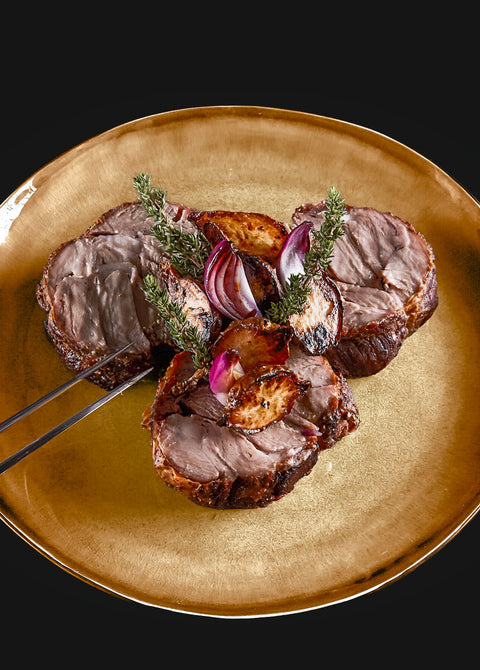 Der Kalbsrollbraten von AUMAERK wird auf einem goldenen Teller mit gerösteten roten Zwiebeln und Kräutern mit einer Servierzange serviert - ein ideales Festtagsessen vor einem markanten schwarzen Hintergrund.